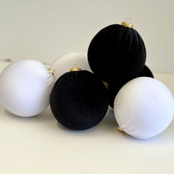 Five velvet Christmas ornaments in black and white with small gold hanging loops, arranged on a light beige surface against a neutral background.