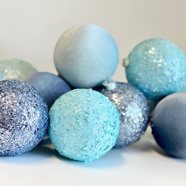 A group of decorative Christmas ornaments in pastel shades of blue, light blue, and silver glitter, arranged on a plain light background.