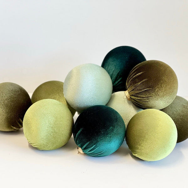 A group of nine velvet Christmas tree ornaments in muted shades of sage green, olive, teal, and pale mint, arranged on a white surface against a white background.