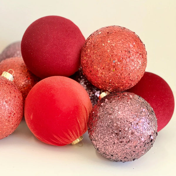 A pile of red and pink Christmas ornaments, including matte velvet balls and glittery sparkly balls, on a light cream background.
