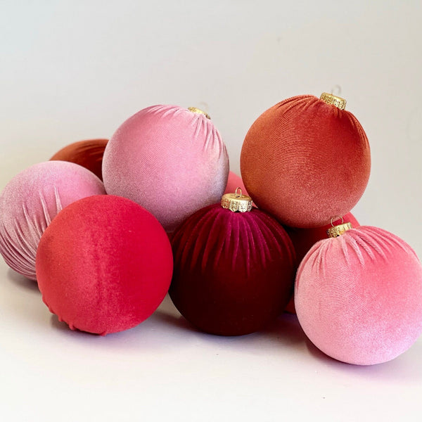 A group of velvet Christmas tree ornaments in various shades of pink, red, and rose gold, arranged on a plain white surface.