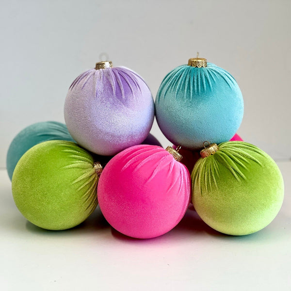 A group of seven velvet Christmas tree ornaments in various bright colors including pink, blue, green, and purple, arranged in a small pile on a white surface.