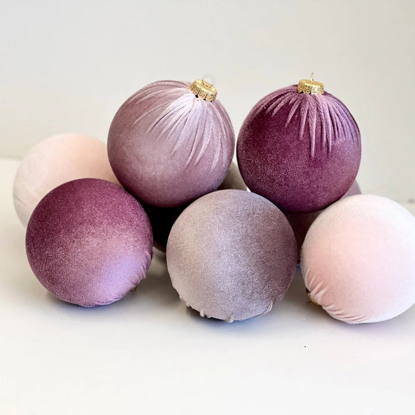Six velvet Christmas ornaments in shades of dusty purple, mauve, and pale pink, arranged on a white surface against a light neutral background.