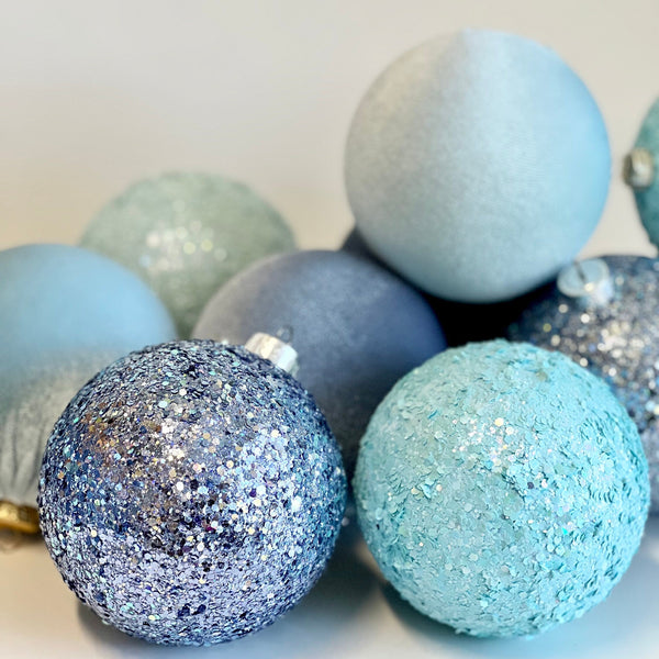 A group of decorative Christmas ornaments in icy blue, light blue, and silver glitter, photographed on a plain light background.