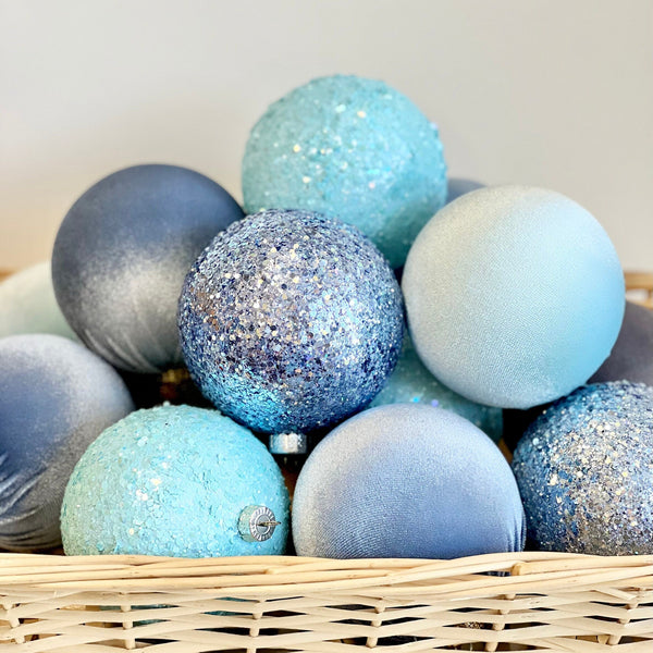 A collection of blue and light blue Christmas ornaments, including matte, glitter, and frosted finishes, arranged in a light brown wicker basket against a neutral beige background.