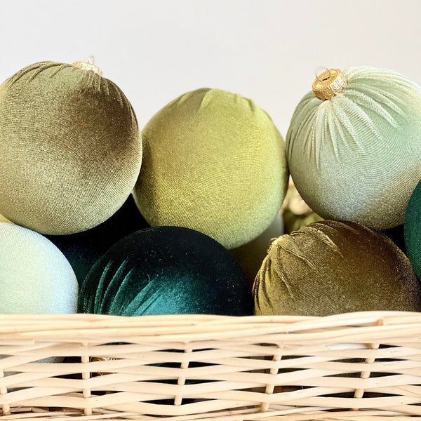 A collection of velvet Christmas ornaments in muted green, sage, and dark teal tones, arranged inside a light natural wicker basket against a clean white background.