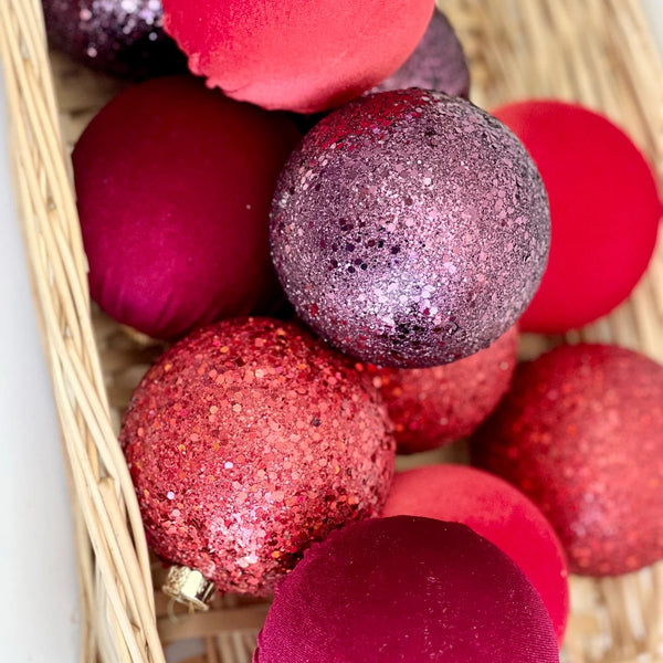 A collection of red and pink Christmas ornaments, including matte velvet, glittery red, and sparkly magenta spheres, arranged in a light beige woven wicker basket.