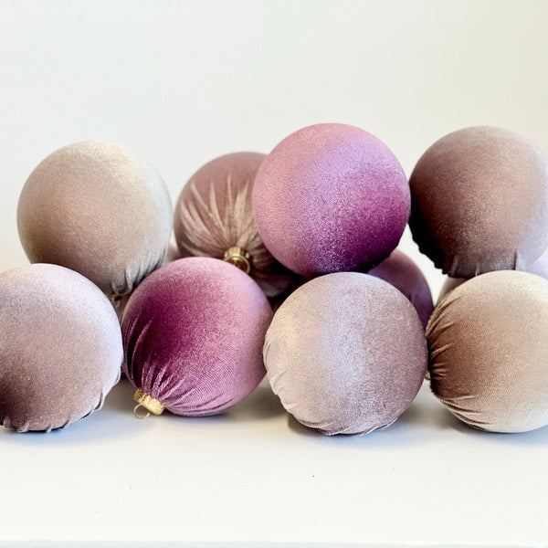 A group of velvet Christmas ornaments in muted shades of mauve, dusty purple, and taupe, arranged on a plain white surface against a light background.