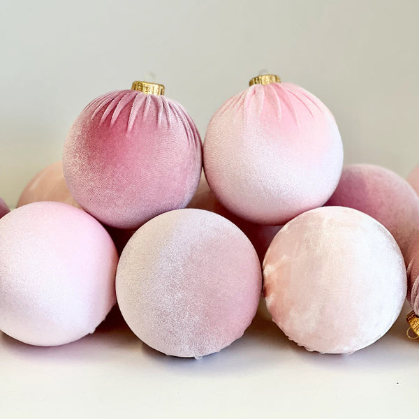A group of soft blush pink velvet Christmas ornaments with gold hanging loops, arranged on a clean white surface against a light neutral background.