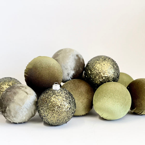 A group of nine decorative Christmas ornaments in muted olive green, sage green, and metallic gold finishes, including textured fabric, velvet, and glitter ball styles, arranged on a plain white surface.