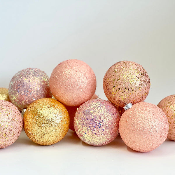 A group of glittery and textured spherical ornaments in pastel pink, peach, and metallic gold, arranged on a plain light background.