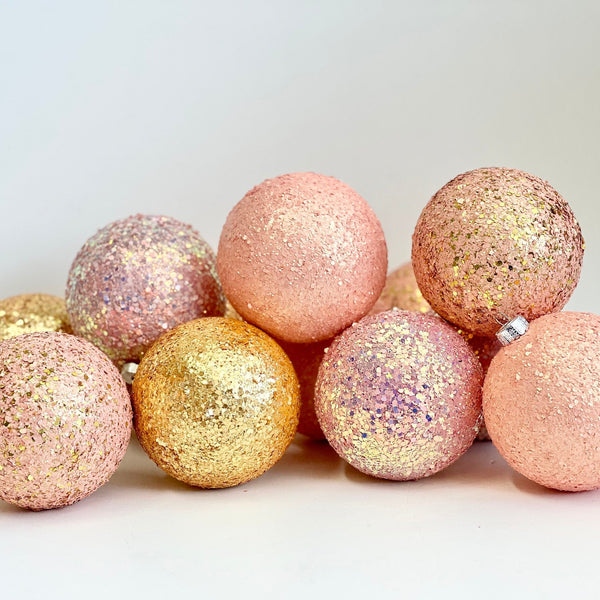 A group of glittery decorative Christmas ornaments in pastel pink, peach, and gold, arranged on a plain white surface against a light gray background.