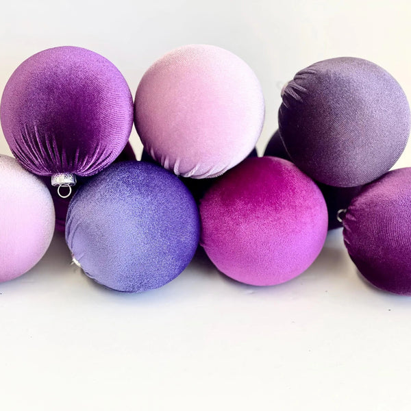 A group of seven velvet Christmas ornaments in various shades of purple, lavender, and pink, arranged on a plain white background.