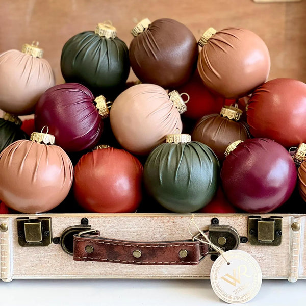 A collection of matte and glossy Christmas ornaments in earthy tones of brown, burgundy, sage green, and dusty pink, arranged inside a vintage-style beige suitcase with a leather handle and a hanging round tag.