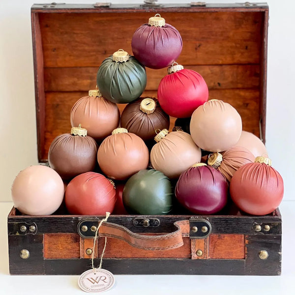 A pyramid arrangement of matte fabric Christmas ornaments in assorted earthy colors, including burgundy, olive green, red, tan, brown, and blush, displayed inside an open vintage wooden suitcase. A hanging tag on the suitcase reads VR.