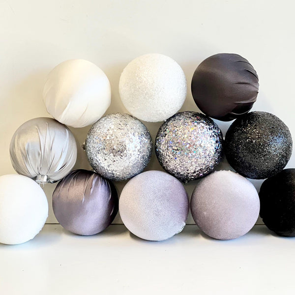 A group of decorative spherical ornaments or balls in assorted finishes and colors, including matte white, glittery silver, iridescent silver, dark charcoal, and soft lavender, arranged against a plain light background.