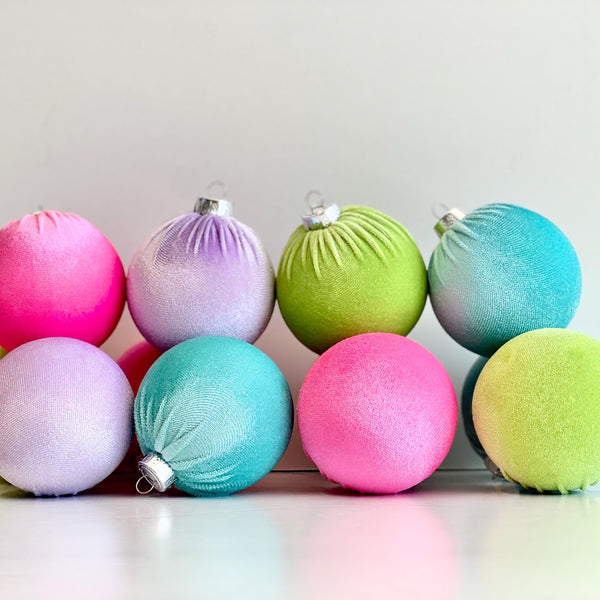 Nine pastel-colored velvet Christmas ornaments in shades of pink, purple, green, and blue, arranged in two rows on a white surface against a light gray background.