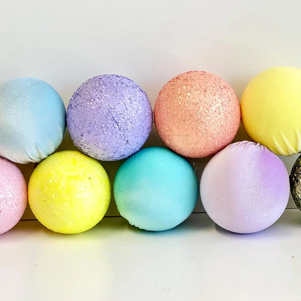 Nine pastel decorative spherical ornaments in various textures, including glitter, fabric, and solid matte finishes, arranged in two rows on a white surface against a white background.