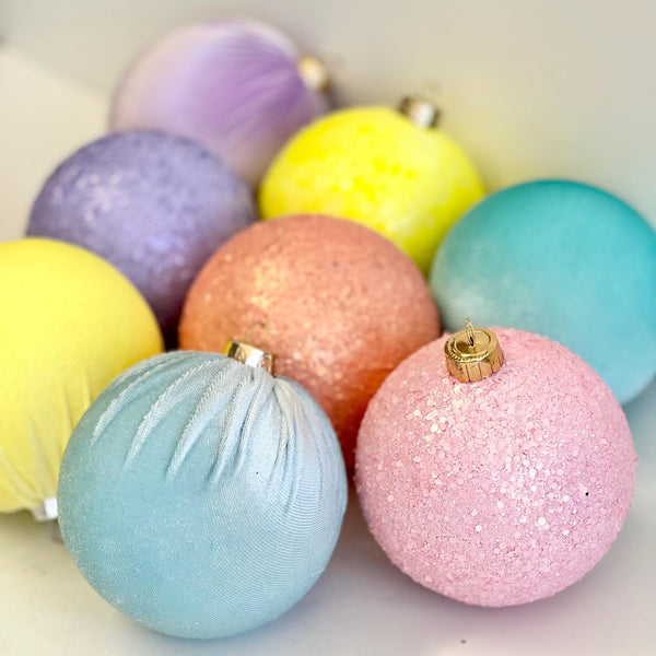 A group of pastel-colored Christmas ornaments in glitter and textured finishes, including pink, light blue, yellow, purple, and peach, arranged on a light surface.