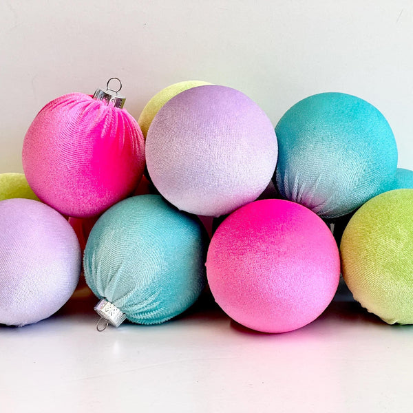 A group of pastel glitter Christmas ornaments in pink, purple, blue, and green, arranged on a white surface against a white background.