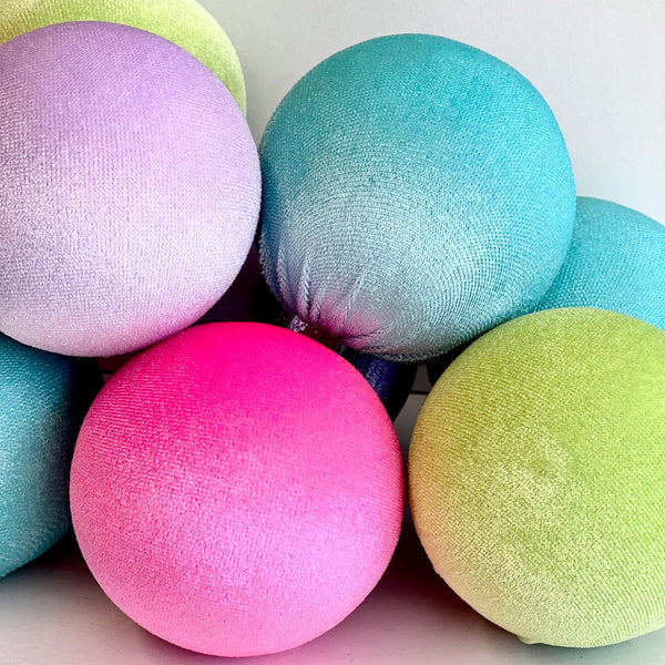A group of round, textured fabric poufs or cushions in pastel and bright colors, including pink, teal, lime green, and lavender, arranged together on a plain light background.