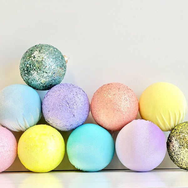 A set of pastel Christmas ornaments arranged in two rows against a plain light background. The ornaments include various colors such as pink, yellow, blue, purple, and green, with some having a glittery texture and others appearing smooth or fabric-textured.