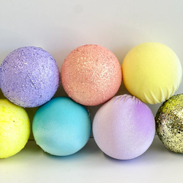 A set of eight decorative bath bombs in various pastel and glitter colors, arranged in two rows on a plain white background.