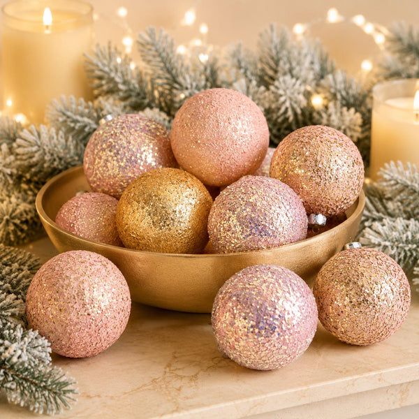 A set of glittery Christmas ornaments in rose gold and gold, with some resting in a gold bowl and others placed in front, styled with frosted pine branches and warm string lights.