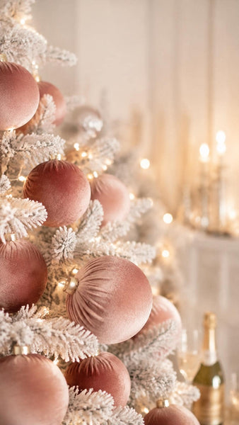 A close-up festive scene of a decorated Christmas tree with large dusty rose velvet ornaments and white frosted branches, softly lit by warm string lights and candles in the background.