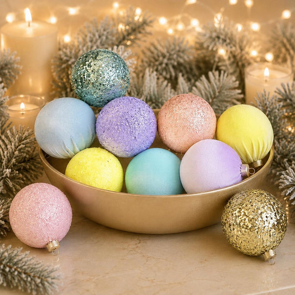 A collection of pastel Christmas ornaments in both matte and glitter finishes, arranged in a gold bowl with decorative candles, pine branches, and fairy lights in the background.