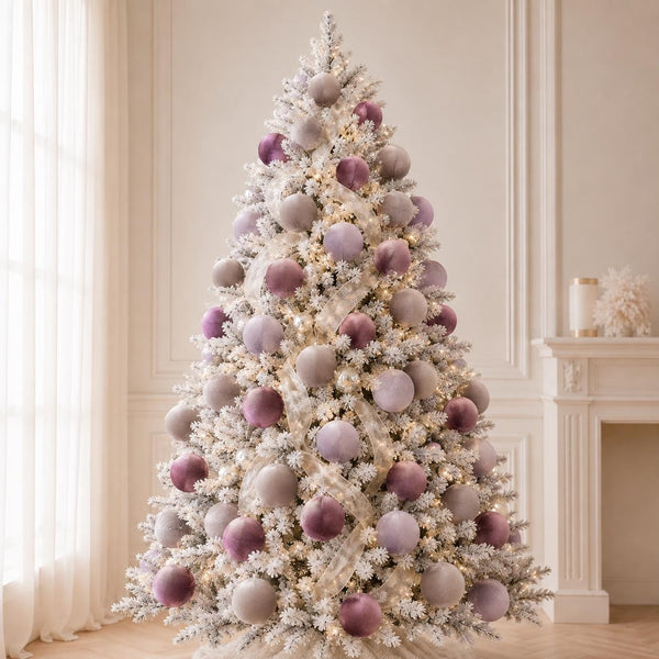 A flocked Christmas tree decorated with white and dusty purple velvet ornaments, lit with warm white string lights, set in a bright, elegant room with a fireplace and curtains.