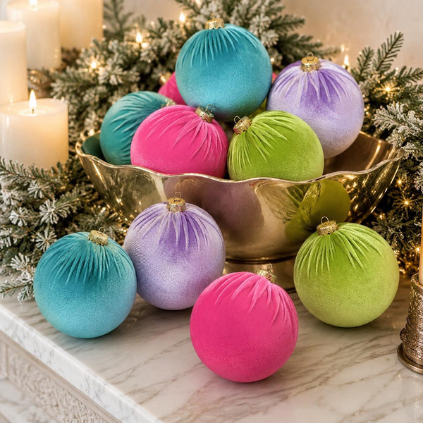 A collection of velvet Christmas ornaments in vibrant shades of turquoise, magenta, lime green, and lavender, arranged in a gold bowl on a marble surface with holiday greenery and candles in the background.