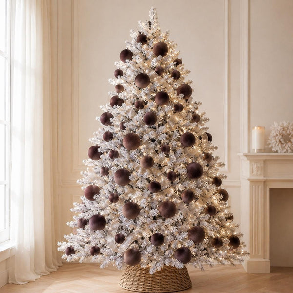 A flocked white Christmas tree with large dark brown ornaments and warm white string lights, displayed indoors in a decorated room with a fireplace and curtains.