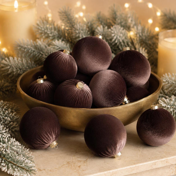 Dark brown velvet Christmas ornaments arranged in a gold bowl with frosted green pine branches and warm string lights in the background.
