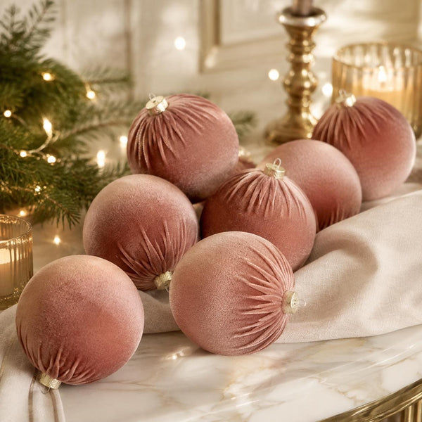 A group of dusty rose velvet Christmas ornaments with gold caps and ribbed texture, arranged on a white marble runner with a white fabric, a lit candle, and a pine tree in the background.