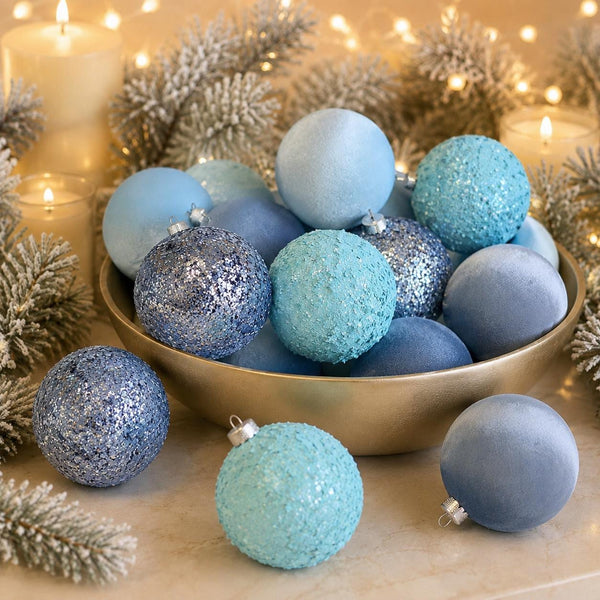 A collection of light blue and turquoise Christmas ornaments, including matte, glitter, and textured finishes, arranged in a gold bowl with festive silver pine branches and warm candlelight in the background.