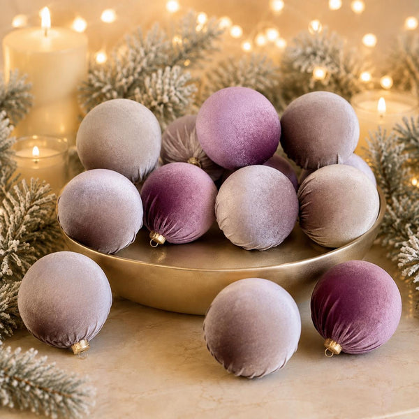 Velvet Christmas ornaments in soft lavender and dusty purple shades, displayed on a gold tray with candles, pine branches, and warm string lights in the background.